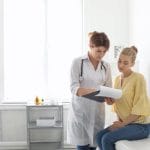 A woman sits on a bench in a doctor's office. The doctor stands next to her, wearing a white coat and holding a clipboard.