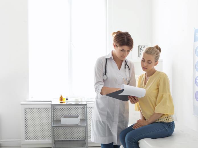 A woman sits on a bench in a doctor's office. The doctor stands next to her, wearing a white coat and holding a clipboard.