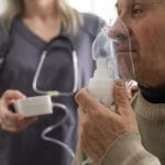 An older man holds an oxygen mask up to his nose and mouth. A nurse stands behind him, holding a portable machine.
