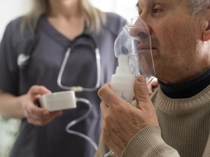An older man holds an oxygen mask up to his nose and mouth. A nurse stands behind him, holding a portable machine.
