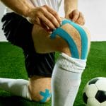 A man wearing cleats and white socks over shin guards is kneeling on turf. He's placing blue kinesiology tape on his knee.