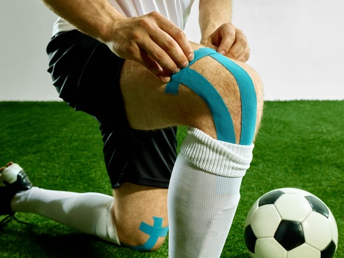 A man wearing cleats and white socks over shin guards is kneeling on turf. He's placing blue kinesiology tape on his knee.