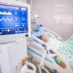A close-up of a medical ventilator machine with a blue screen and yellow text. Someone is hooked up to the machine.