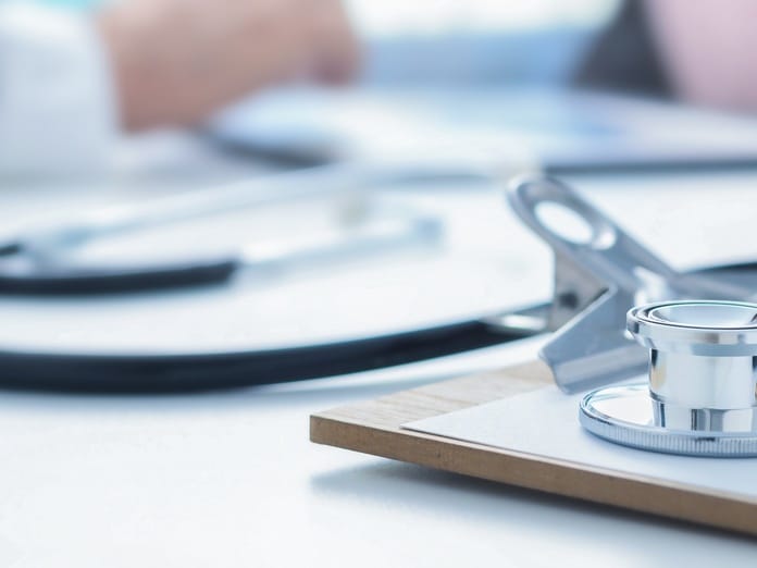A close-up view shows a stethoscope sitting on top of a brown clipboard. A pen sits on the clipboard, too.