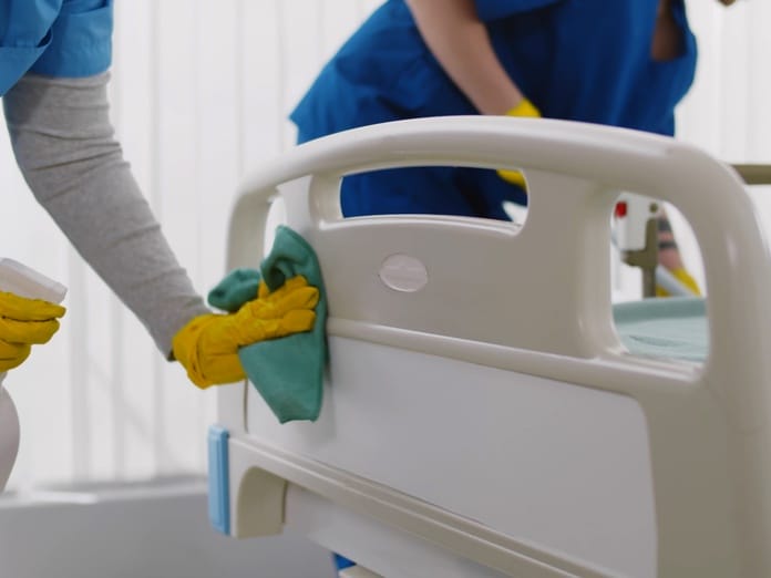 A close view of two workers in blue scrubs cleaning a hospital bed rail with disinfectant spray and a cloth.