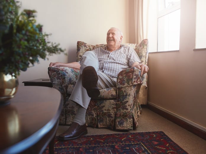 An elderly man smiles while sitting in an armchair beside two large windows, with sunlight pouring into the room.